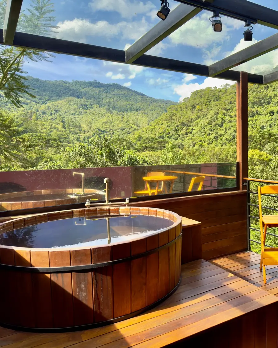 Deck do Studio Lua Nova com ofurô de madeira e mesa, vista para as montanhas sob pérgola de vidro