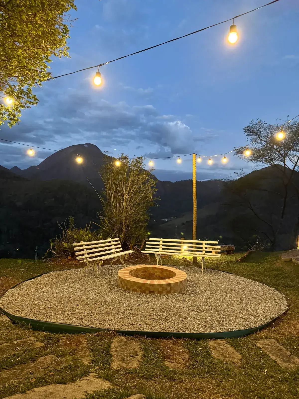 Roda de fogueira ao entardecer no Refúgio Céu Azul, com bancos brancos, luzes penduradas e as montanhas de Itaipava ao fundo