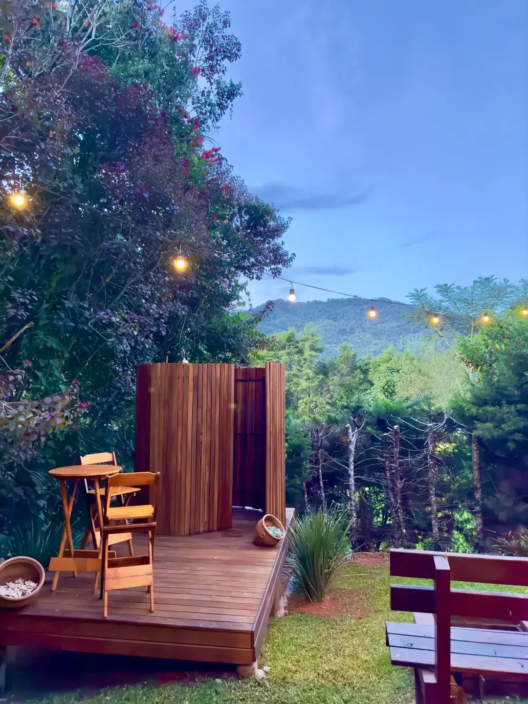 Cabine de ducha externa em madeira no jardim do Chalé, ao entardecer com luzes acesas