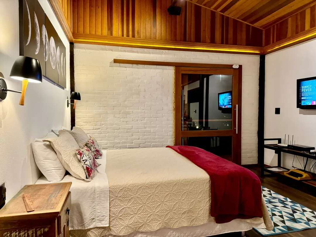 Quarto do Chalé com parede de tijolinho branco, cama de casal e roupa de cama clara
