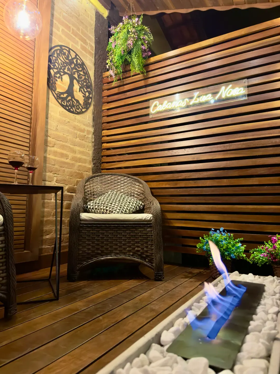 Deck do Chalé à noite com poltronas de vime, mesa lateral com taças de vinho e letreiro Cabanas Lua Nova aceso
