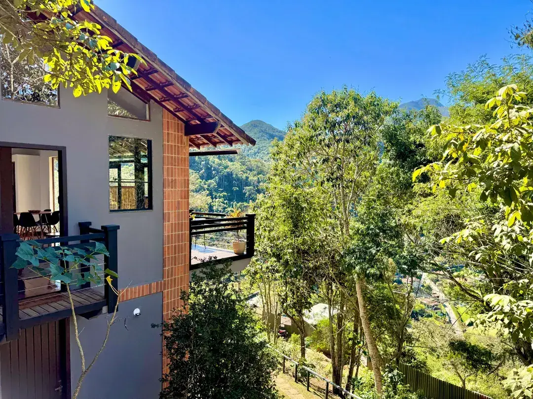 Fachada da Cabana Orion entre as árvores, com as montanhas de Itaipava ao fundo em dia claro