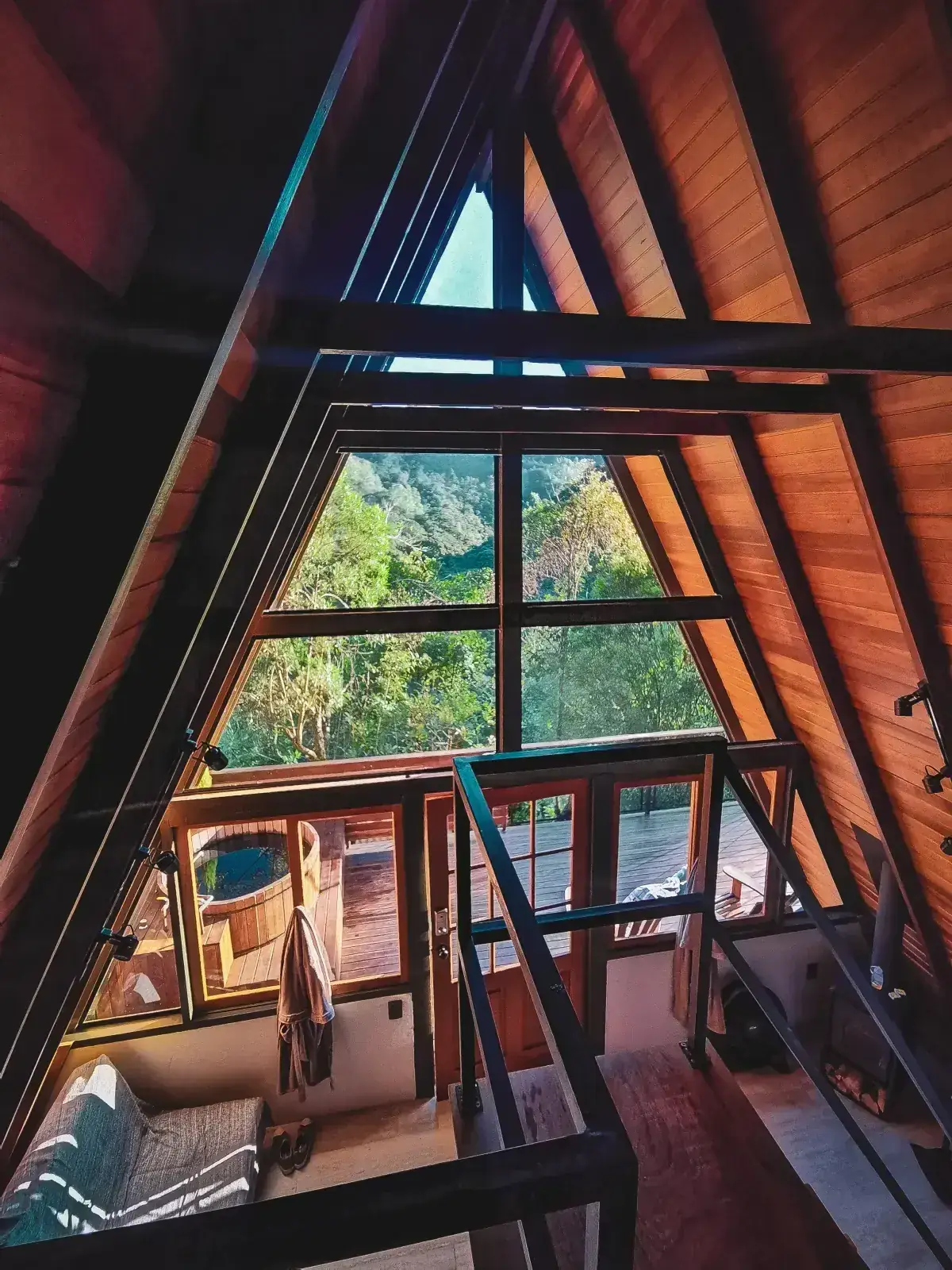 Interior da Cabana Solaris visto do mezanino: a parede triangular de vidro do A-frame emoldurando a mata