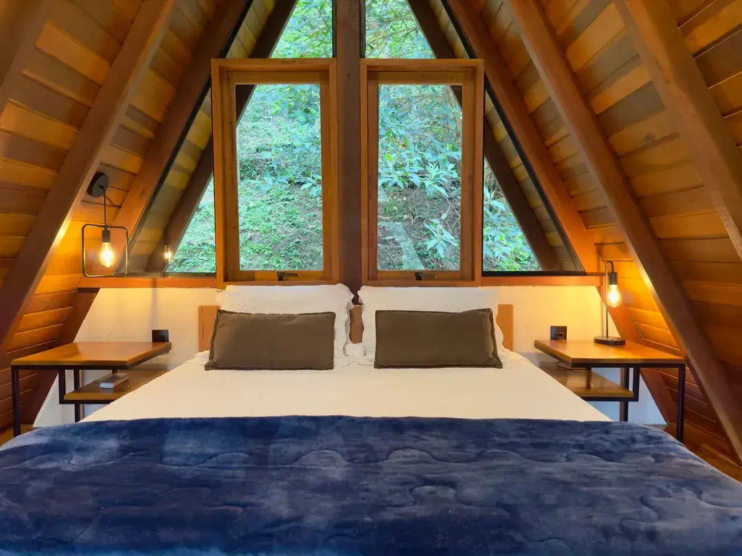 Cama no mezanino sob o telhado A-frame, com janelas emoldurando o verde
